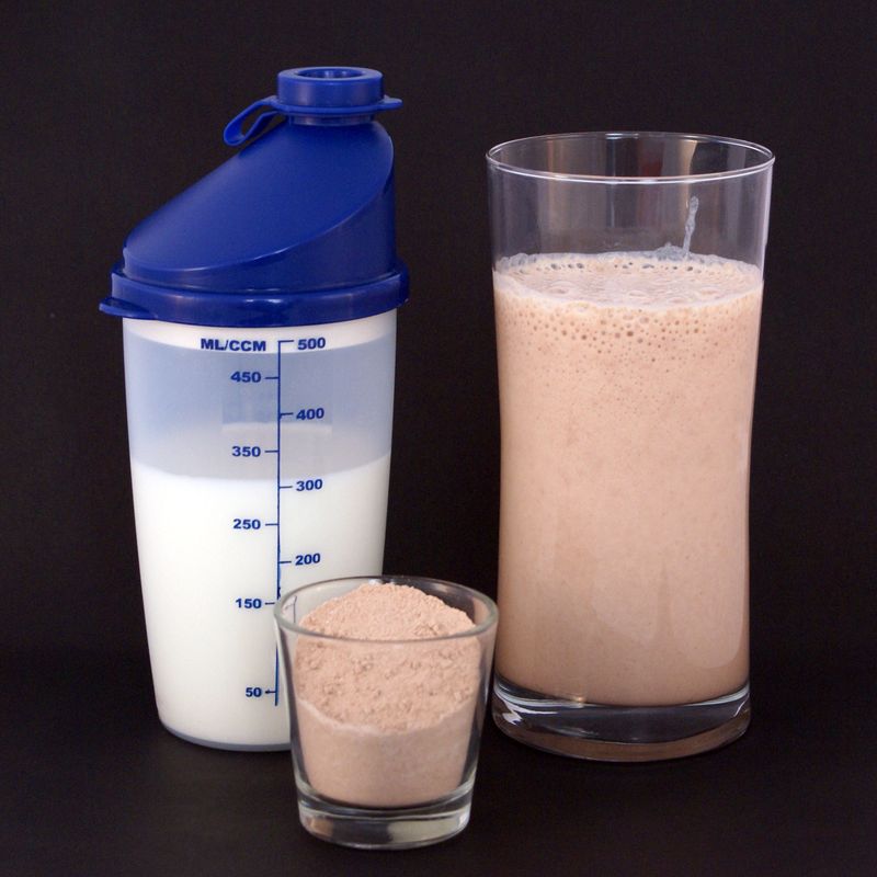 Protein powder in a glass beside a shaker bottle of milk and a finished shake